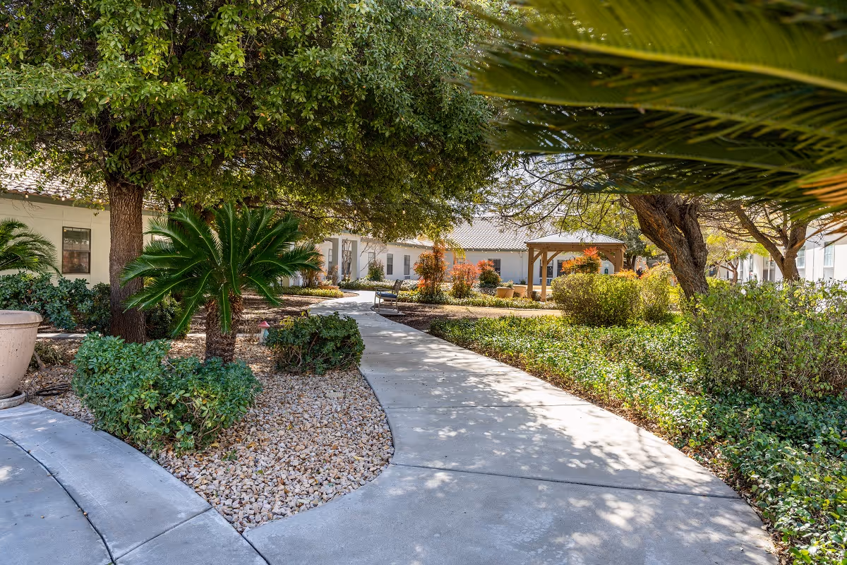 A landscaped outdoor garden area at Inspirations of River Centre featuring a curved concrete pathway surrounded by green shrubs, small palm trees, and other plants. There is a wooden gazebo in the background and white buildings with windows surrounding the garden.