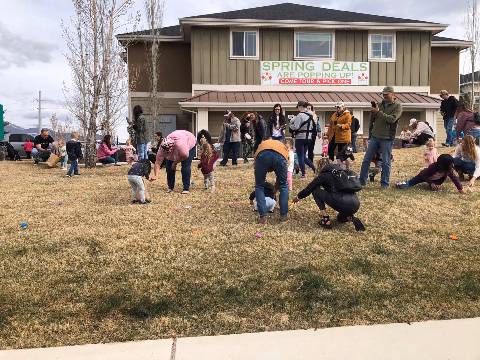A group of adults and children participating in an outdoor Easter egg hunt on a grassy area in front of a two-story building. The building has a sign that reads 'SPRING DEALS ARE POPPING UP! COME TOUR & PICK ONE'. The sky is cloudy and the participants are scattered across the lawn picking up colorful eggs.