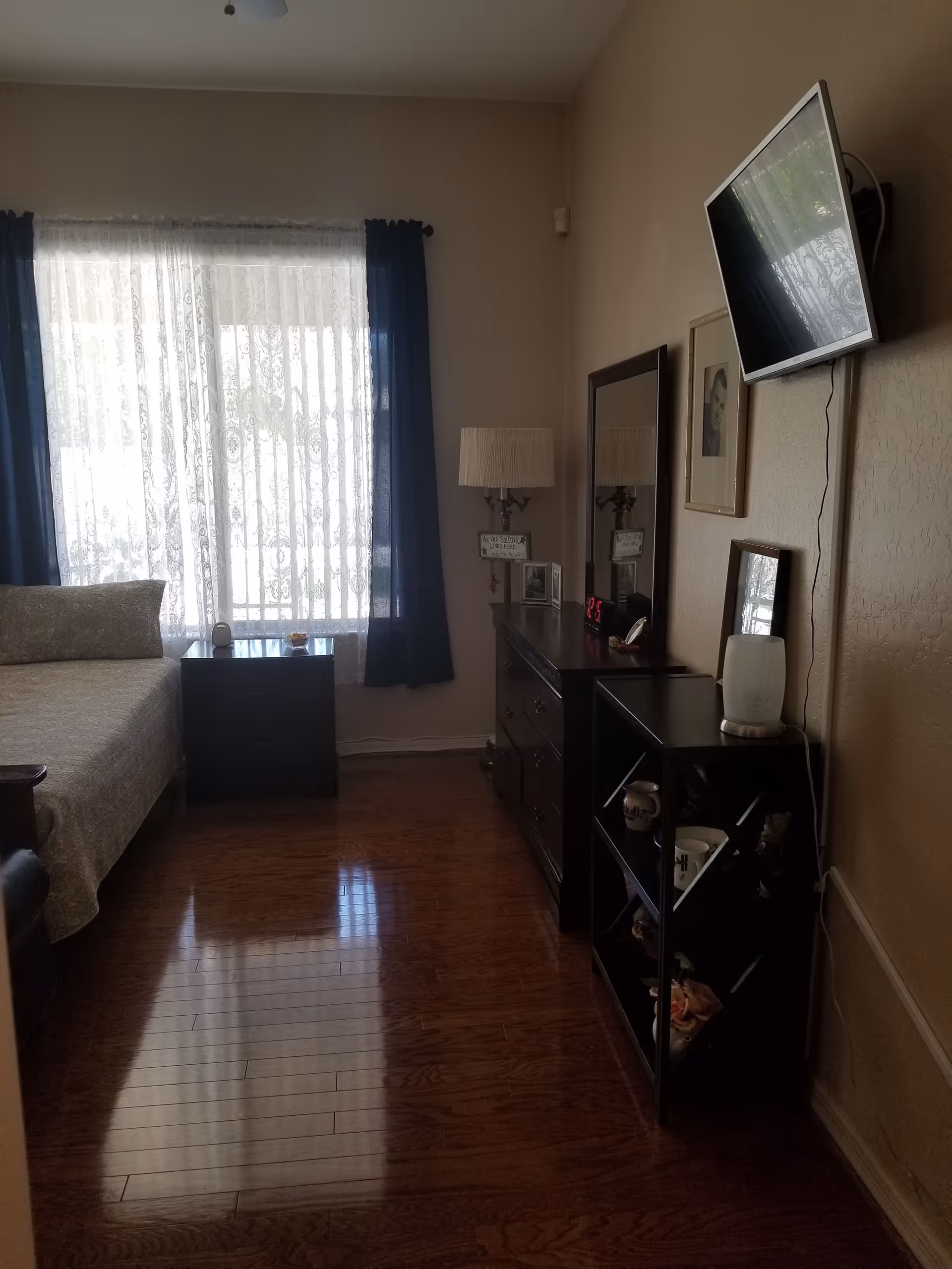 A bedroom with a bed on the left side covered with a patterned bedspread. Next to the bed is a dark wooden nightstand with a small device and a bowl on top. A window with sheer white curtains and dark blue drapes lets in natural light. On the right side of the room, there is a dark wooden dresser with a mirror above it, a lamp, framed pictures, and a digital clock showing 2:25. Above the dresser, a flat-screen TV is mounted on the wall. The floor is polished wood.