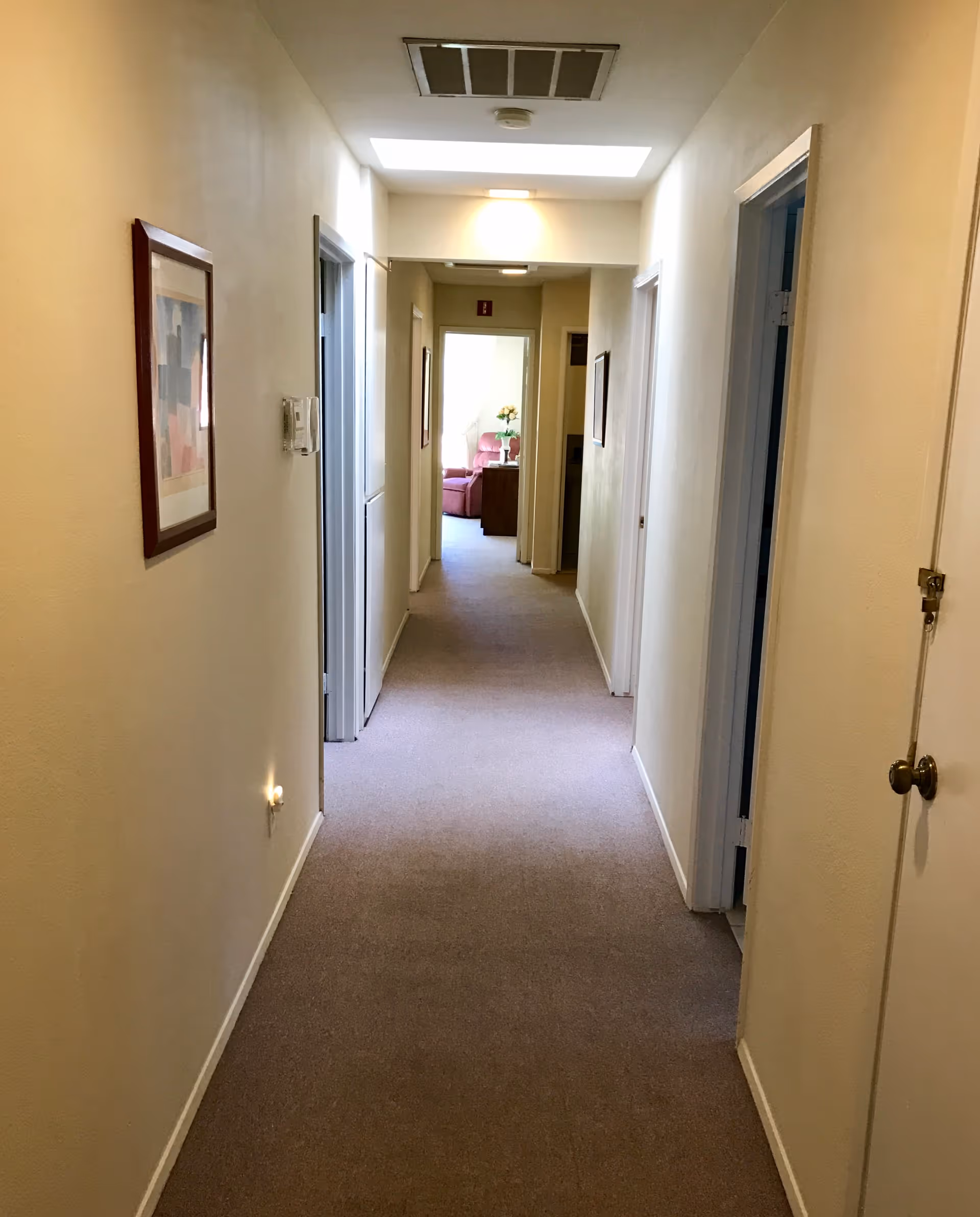 A long, narrow hallway in a residential care facility with beige walls and carpeted floor. Several doorways line both sides of the hallway, and framed artwork hangs on the left wall. At the end of the hallway, a room is visible with a pink armchair, a small table, and a vase with flowers.