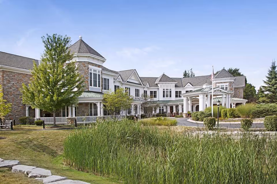 Exterior view of a large, elegant senior living facility building with stone and white siding, multiple windows, a covered entrance with columns, surrounded by greenery including trees, bushes, and tall grass under a clear blue sky.