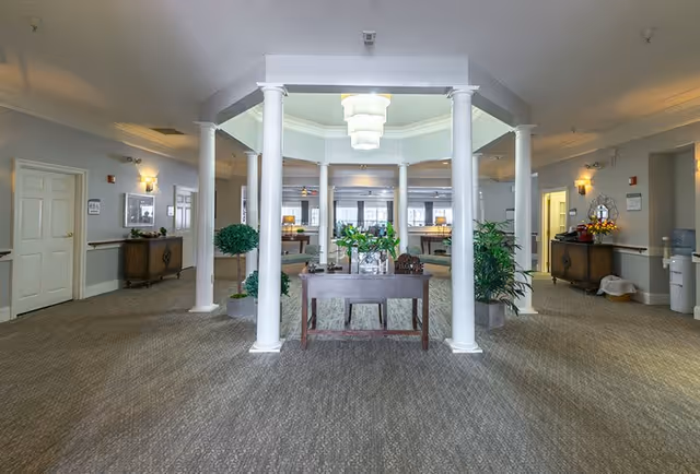 A spacious and well-lit common area with a central wooden table adorned with plants and decorative items. The room features white columns supporting a ceiling with a chandelier, carpeted floors, and walls painted in a soft neutral color. There are multiple doors, side tables with lamps, plants, and a water dispenser visible in the background.