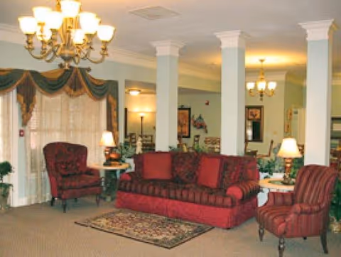 A cozy living room area with a red sofa and two matching armchairs, a small patterned rug on the carpeted floor, white columns, table lamps, and chandeliers providing warm lighting. The room features large windows with draped curtains and decorative valances.