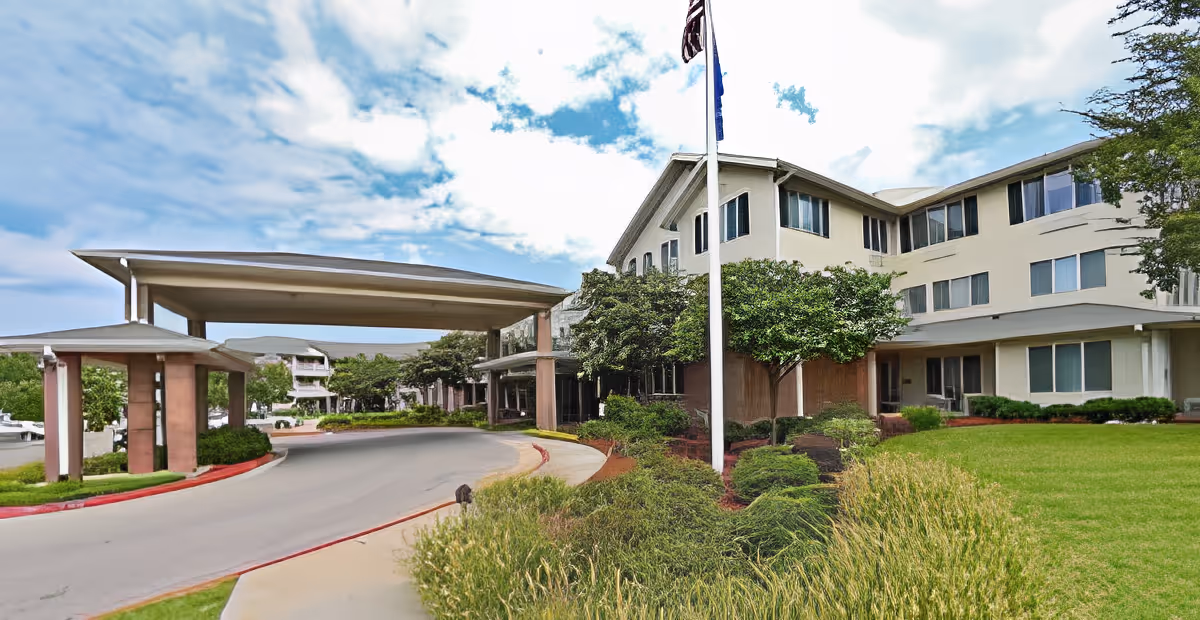 Exterior view of Morada Broken Arrow facility showing a multi-story building with numerous windows, a covered driveway entrance, well-maintained landscaping with bushes and grass, and a flagpole with an American flag against a partly cloudy sky.