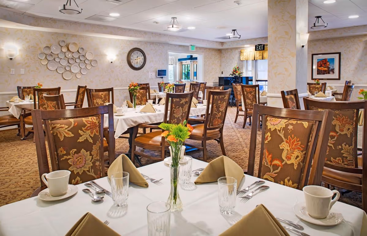 A well-lit dining room with multiple tables set with white tablecloths, beige folded napkins, glassware, cups, and silverware. Each table has a small vase with flowers. The room features floral-patterned chairs, a wall clock, decorative plates on the wall, and a piano near the exit door.