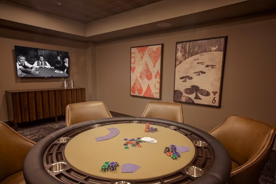 A cozy room with a round poker table in the center, surrounded by four leather chairs. The table has poker chips and playing cards on it. On the wall behind the table, there are two large framed artworks featuring playing card suits. A flat-screen TV mounted on the wall shows a black and white image of people playing poker. Below the TV is a wooden cabinet with glassware on top.