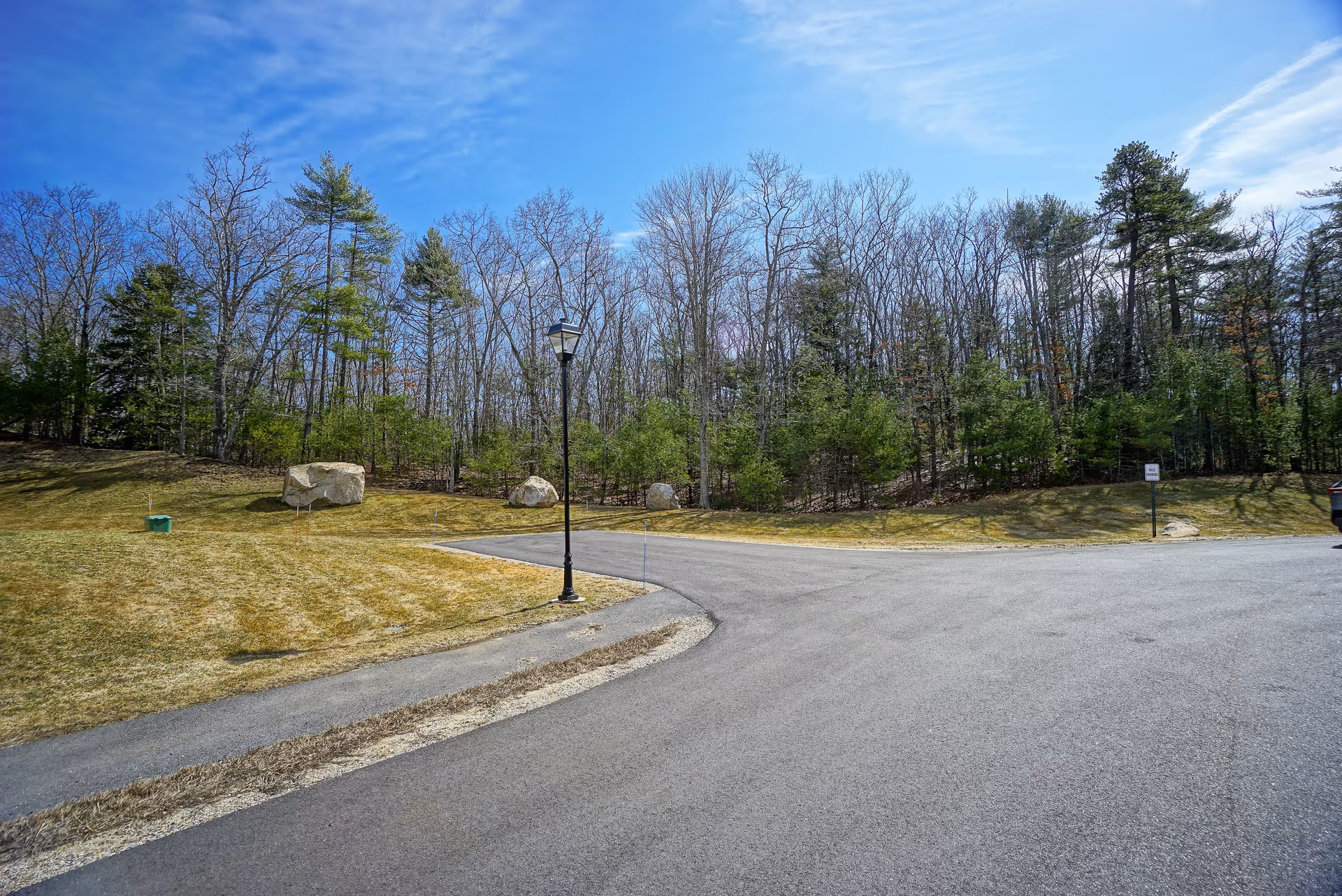 A paved road curves through an outdoor area with a grassy lawn, a few large rocks, a lamppost, and a backdrop of leafless and evergreen trees under a blue sky with wispy clouds.