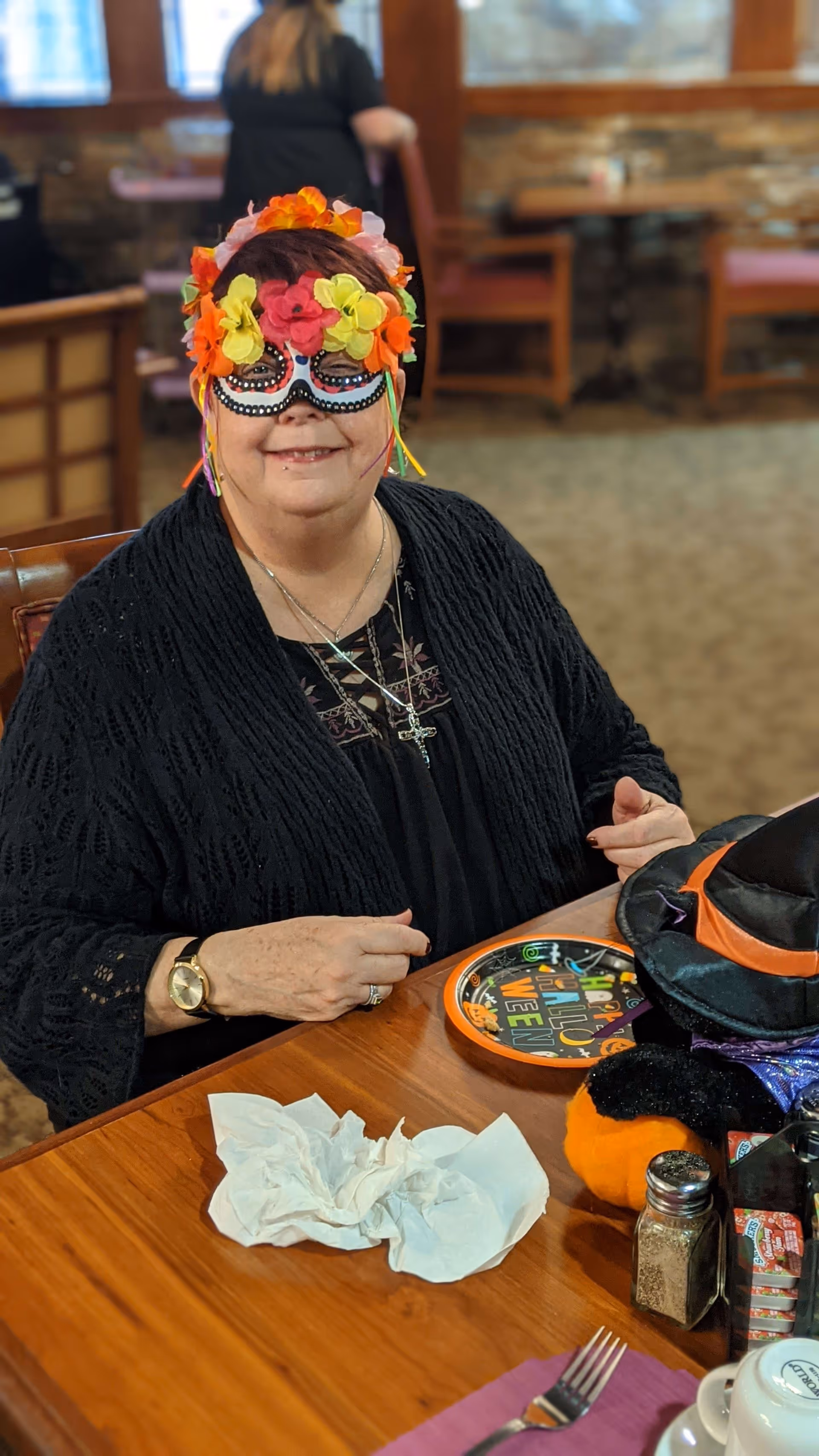 A woman wearing a colorful floral mask and black clothing sits at a wooden table in a dining area decorated for Halloween, with a Halloween-themed plate, a small pumpkin, and a witch hat on the table.