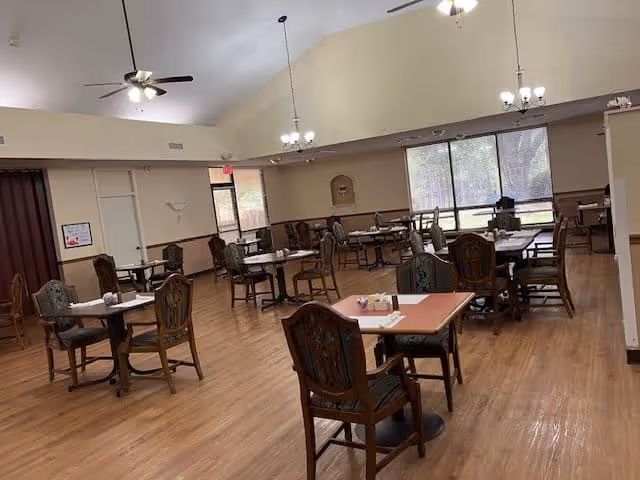 A spacious dining room with multiple square tables and wooden chairs arranged neatly. The room has a high ceiling with ceiling fans and hanging light fixtures. Large windows on one side allow natural light to enter, and the floor is wooden.