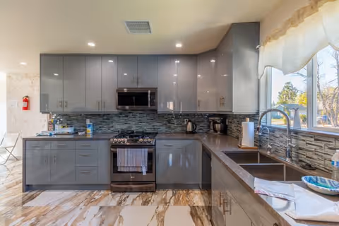 Modern gray kitchen with glossy cabinets, stainless steel stove and microwave, tiled backsplash, double sink under a window, and marble-patterned floor.