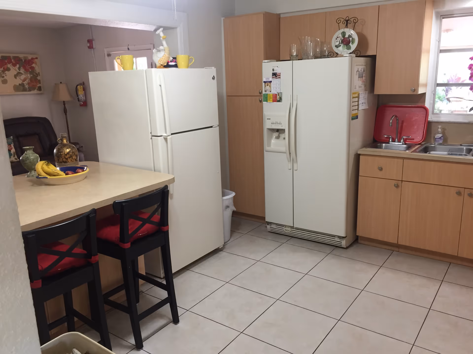 A kitchen area with two white refrigerators, wooden cabinets, a countertop with two black chairs with red cushions, a bowl of bananas and apples, and a sink with a red dish rack and hand soap near a window.