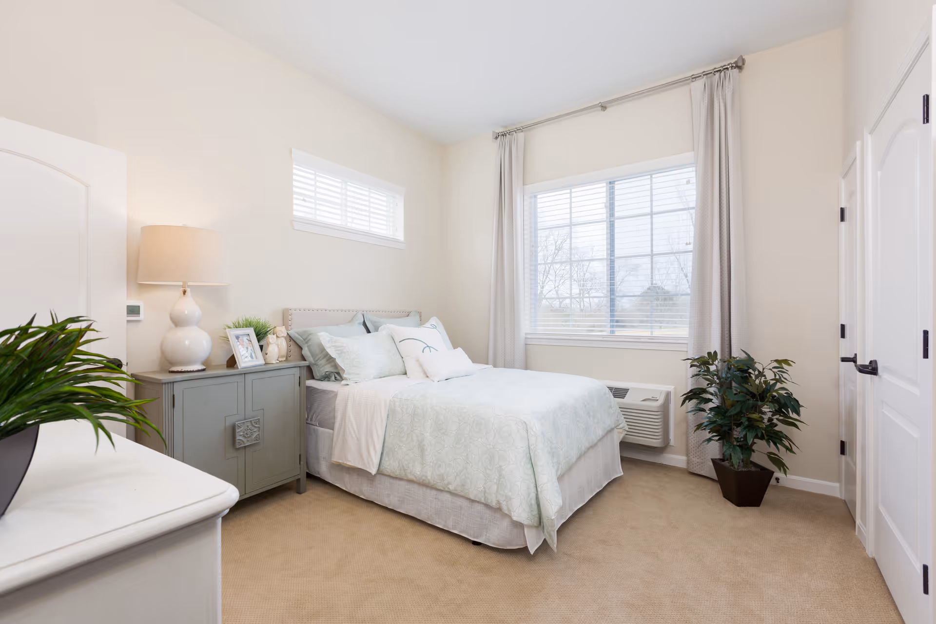 A bright and clean bedroom with a neatly made bed featuring light-colored bedding. There is a small gray nightstand with a white lamp, framed photos, and a small plant. The room has beige carpet, a large window with white blinds and light curtains, a smaller window above the bed, and a potted plant near the window. The walls are painted a soft cream color.