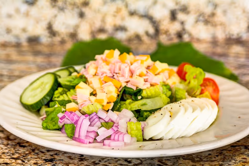 A plate of fresh salad containing sliced cucumbers, chopped red onions, diced ham and cheese, lettuce, cherry tomatoes, and sliced hard-boiled eggs, placed on a granite countertop.