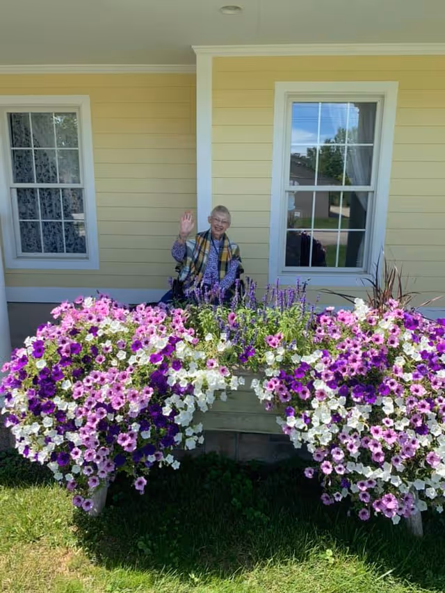 An elderly woman sitting in a wheelchair on a porch in front of a yellow house with white trim, waving and smiling. In front of her is a large planter overflowing with vibrant purple, pink, and white flowers. Two windows with white frames are visible behind her.