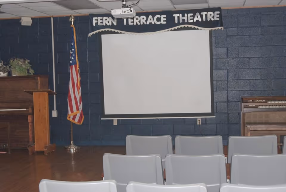 Interior view of a small theater room with a white projection screen mounted on a dark blue brick wall. Above the screen, there is a sign that reads 'FERN TERRACE THEATRE'. In front of the screen, there are several rows of gray chairs. To the left of the screen, there is an American flag on a stand and a wooden podium next to a piano. To the right of the screen, there is an organ.