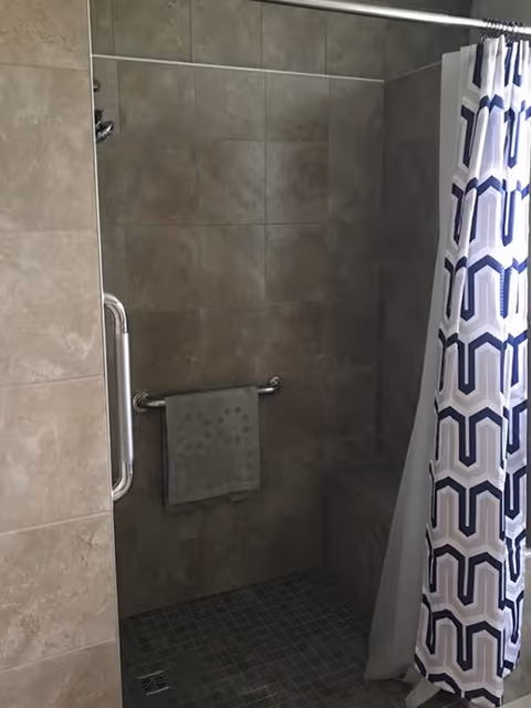 Tiled walk-in shower with a grab bar, small built-in bench, towel on a rail, and a patterned shower curtain.