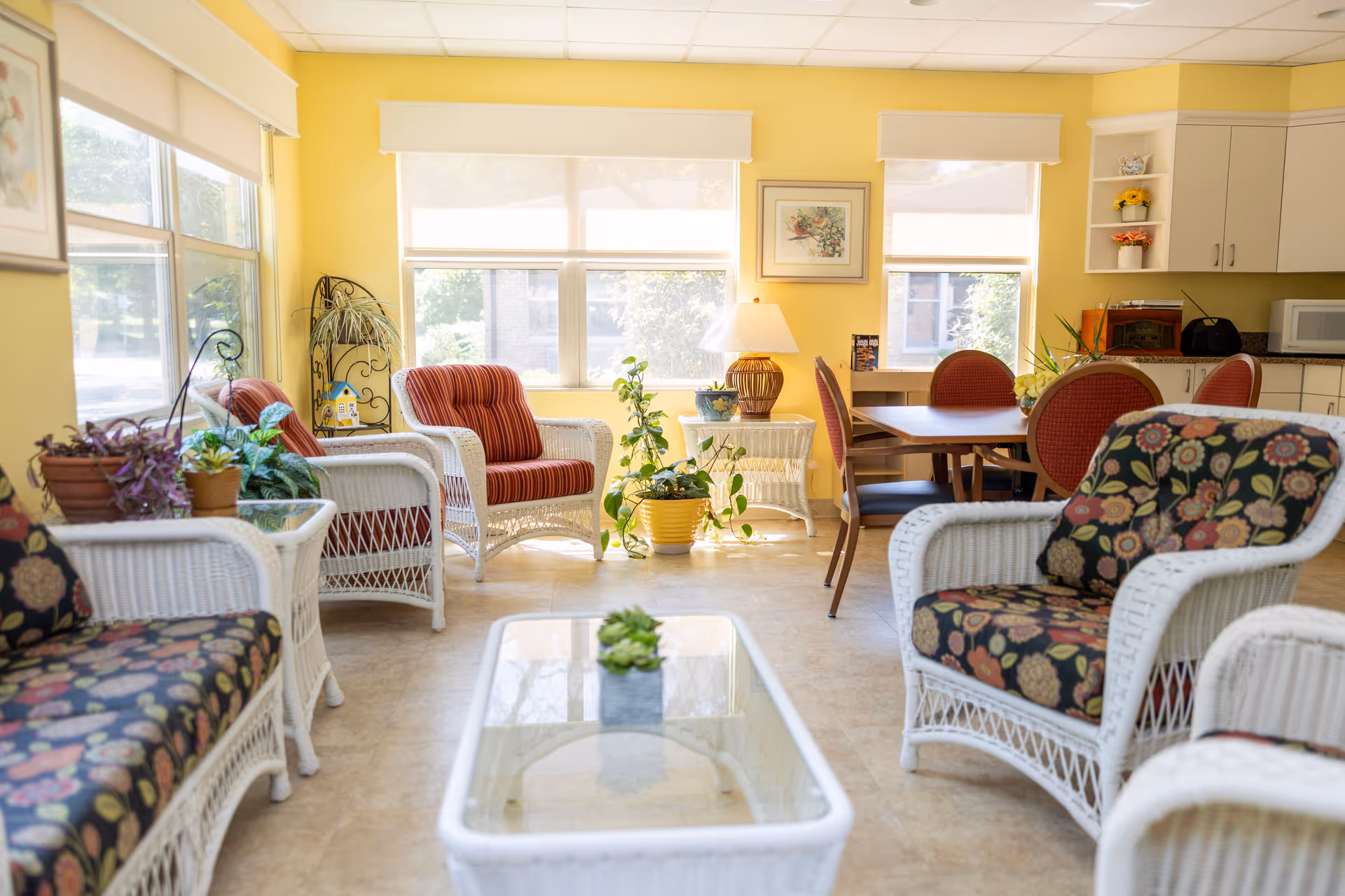 Bright communal living room with white wicker chairs, patterned cushions, potted plants, a glass-top coffee table, and a small dining area.