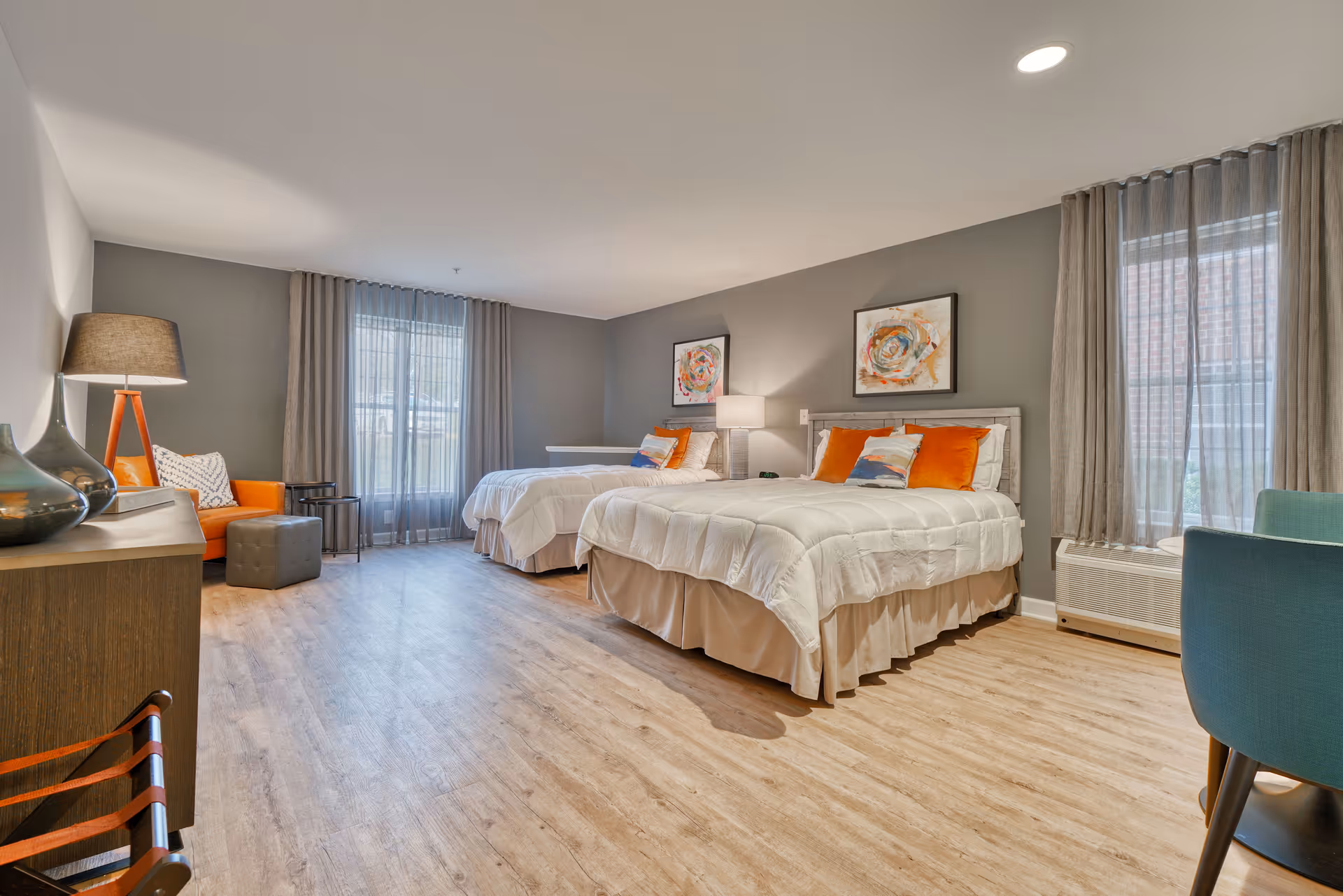 A spacious bedroom with two beds, each with white bedding and orange accent pillows. The room features light wood flooring, gray walls, two large windows with sheer curtains, a wooden dresser with decorative vases, an orange armchair with a floor lamp, and a small round table with a teal chair.