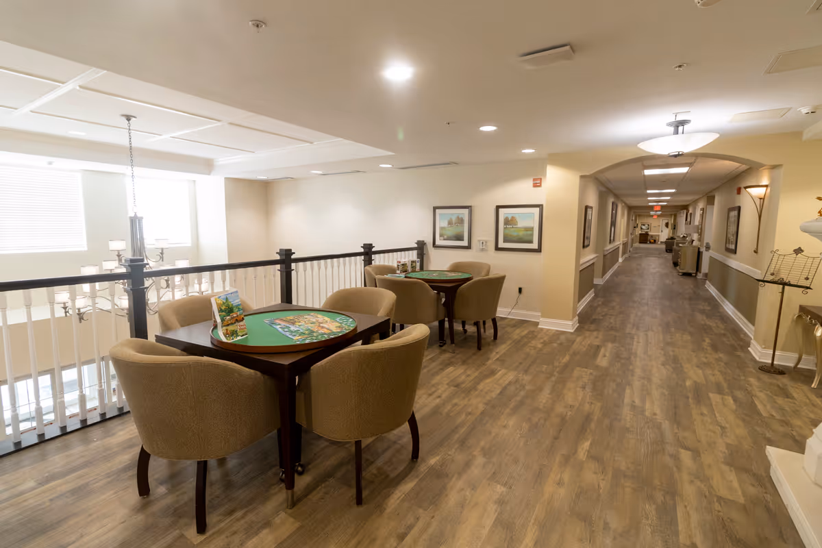 Interior view of a senior living facility hallway with two small tables and four chairs each, set up for games or activities. The space has wood flooring, beige walls, framed artwork, and overhead lighting. A railing overlooks a lower level with a chandelier visible through the window.