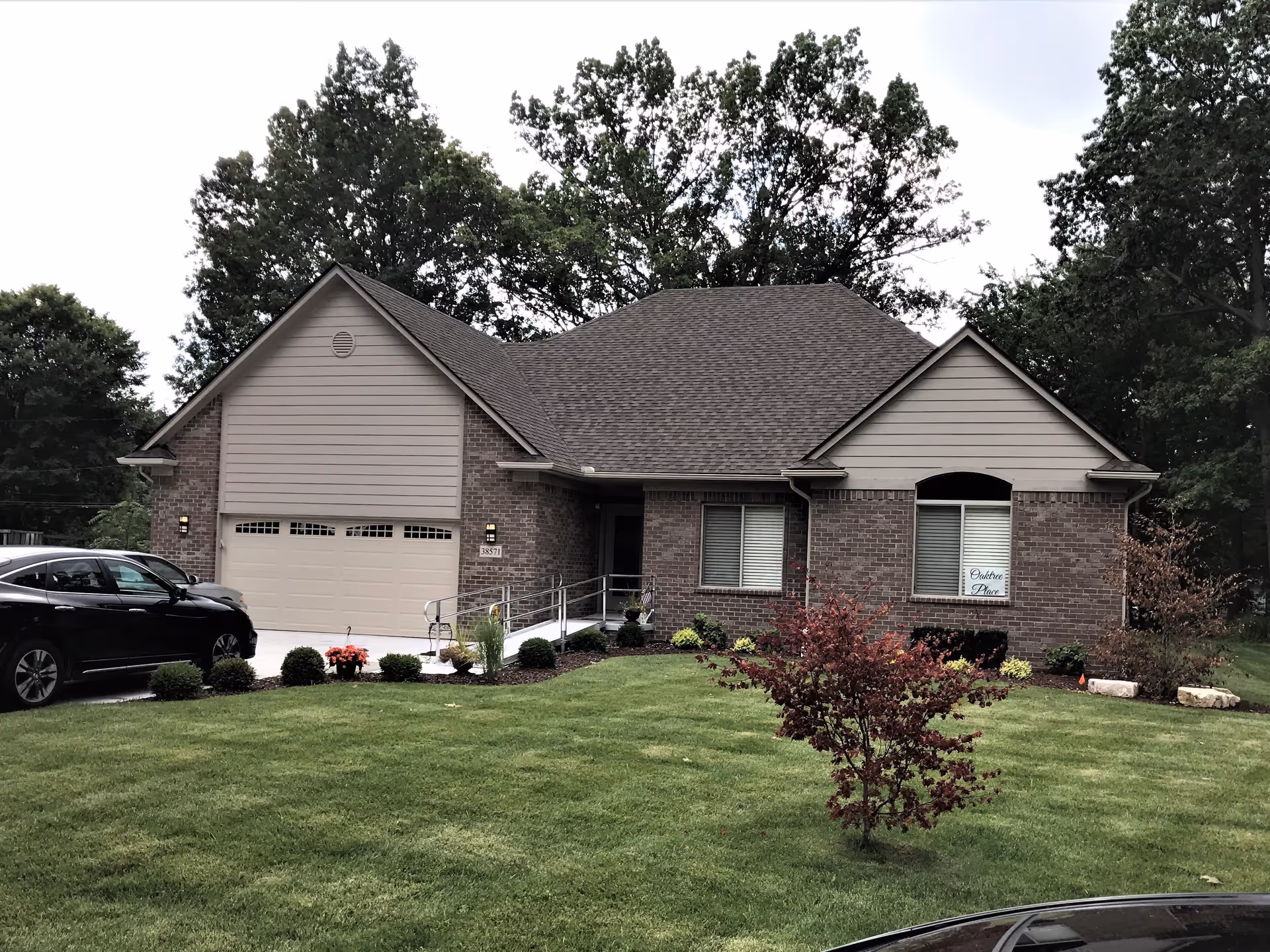 Front exterior view of a single-story brick house with a two-car garage, a small ramp leading to the front door, manicured lawn, small bushes, and a tree with red leaves in the yard. There are two cars parked in the driveway and trees in the background.
