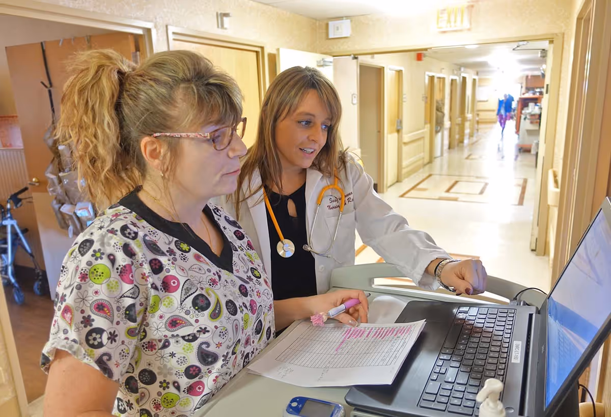 Two healthcare workers in a nursing facility hallway looking at a laptop on a mobile workstation. One is wearing a white coat with a stethoscope and the other is in patterned scrubs holding a paper with highlighted text. The hallway has several doors and an exit sign is visible.