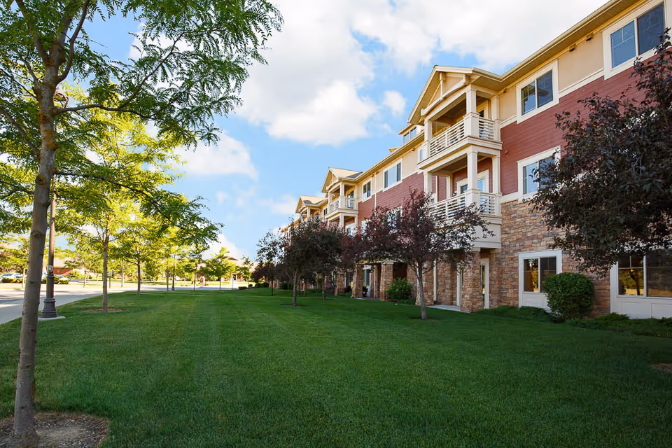 Exterior view of a multi-story senior living facility building with red and beige siding and stone accents. The building has balconies and is surrounded by a well-maintained green lawn with several trees. The sky is partly cloudy with blue patches.