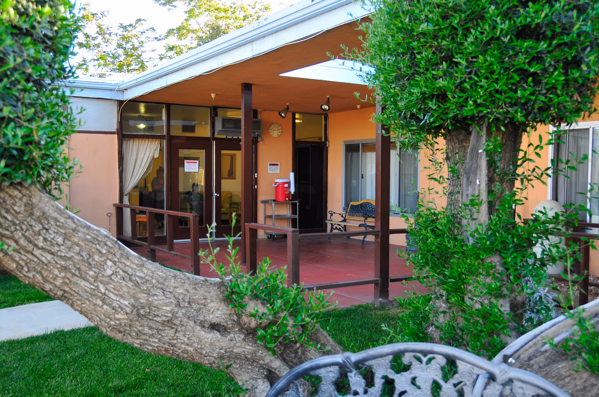 Covered patio entrance to a senior living facility with glass doors, benches, and trees in the foreground.