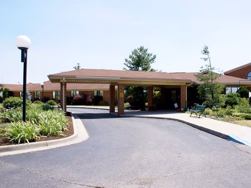 Driveway and covered entrance/portico of a single-story brick senior living facility with landscaped beds, a lamppost, and a bench.