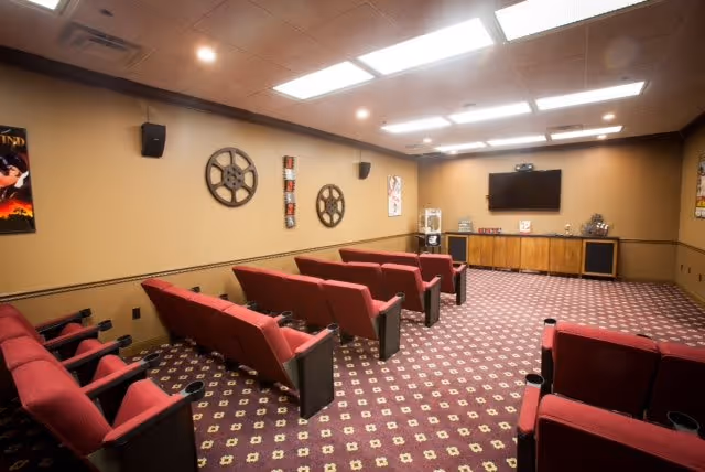 A small theater room with rows of red cushioned seats facing a large flat-screen TV mounted on a beige wall. The room has patterned carpet flooring and movie-themed decorations including film reels and posters on the walls. There is a popcorn machine and speakers at the front of the room.