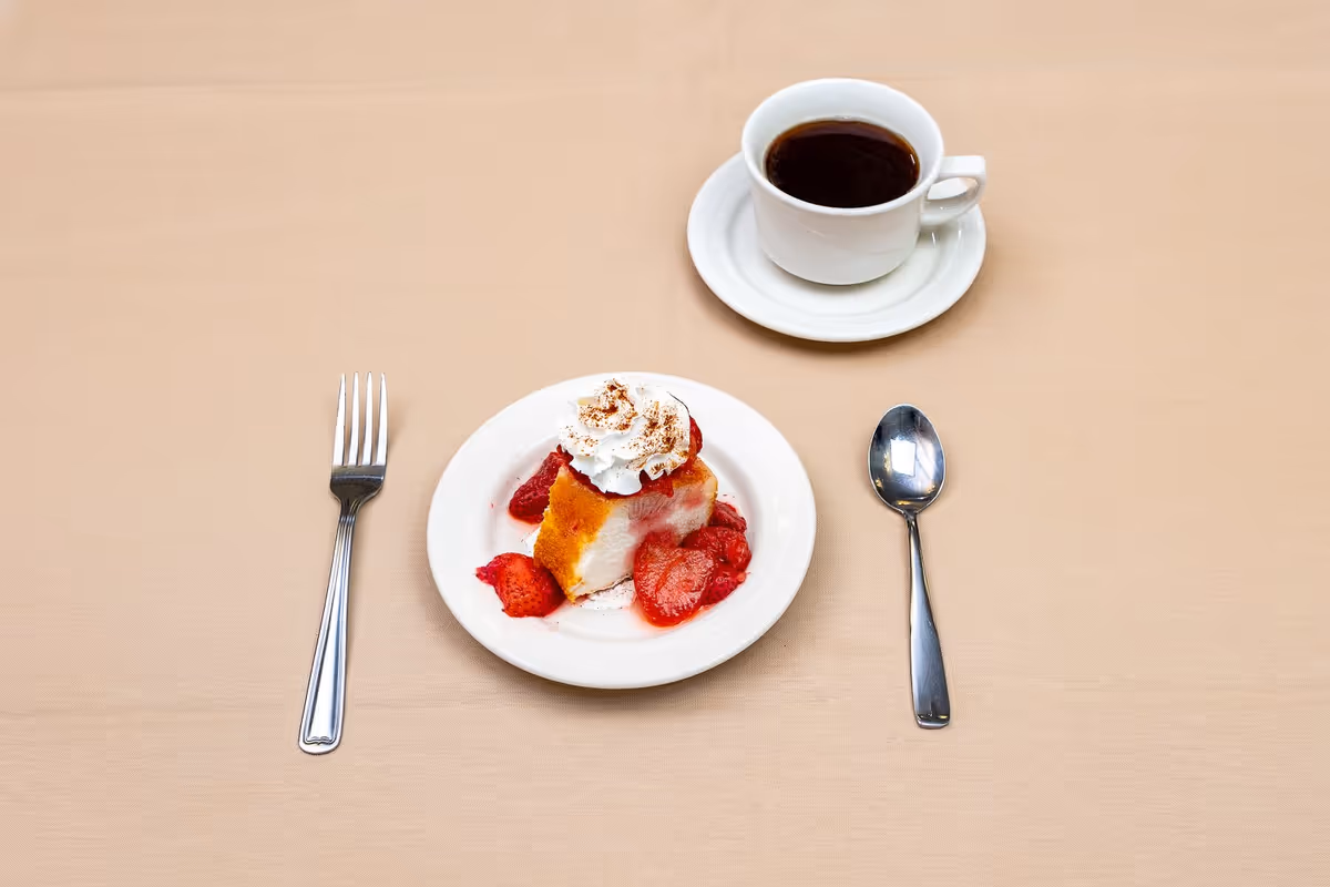 A dessert plate with a slice of cake topped with whipped cream and strawberries, placed on a beige tablecloth. To the left of the plate is a fork, and to the right is a spoon. Behind the plate is a white cup filled with black coffee on a matching saucer.