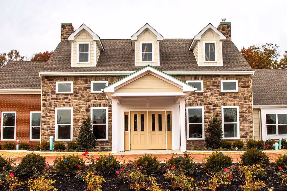 Front exterior view of Mullica Gardens Assisted Living facility showing a large building with a stone and brick facade, multiple windows, a central entrance with double doors under a small covered porch, and landscaped bushes and flowers in the foreground.