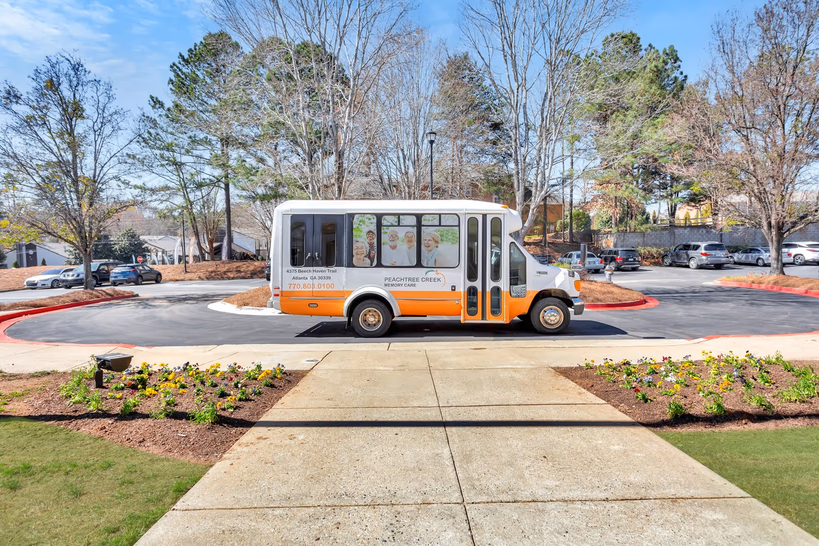 A white and orange shuttle bus parked on a driveway in front of a landscaped area with flowers and trees. The bus has images of smiling elderly people on its windows and displays the name Peachtree Creek Memory Care along with contact information and address in Atlanta, Georgia.