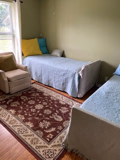 A small bedroom with two single beds covered in light blue bedspreads, each with colorful pillows. There is a beige armchair with a yellow cushion near a window with white curtains. A patterned red and beige rug covers part of the wooden floor.