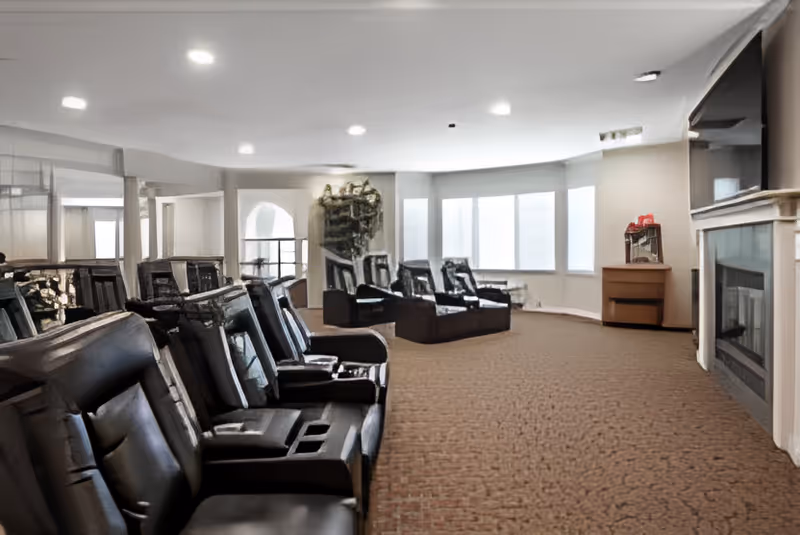 A spacious lounge area with multiple black leather reclining chairs arranged in rows facing a fireplace and a large flat-screen TV mounted above it. The room has a carpeted floor, large windows letting in natural light, and neutral-colored walls.