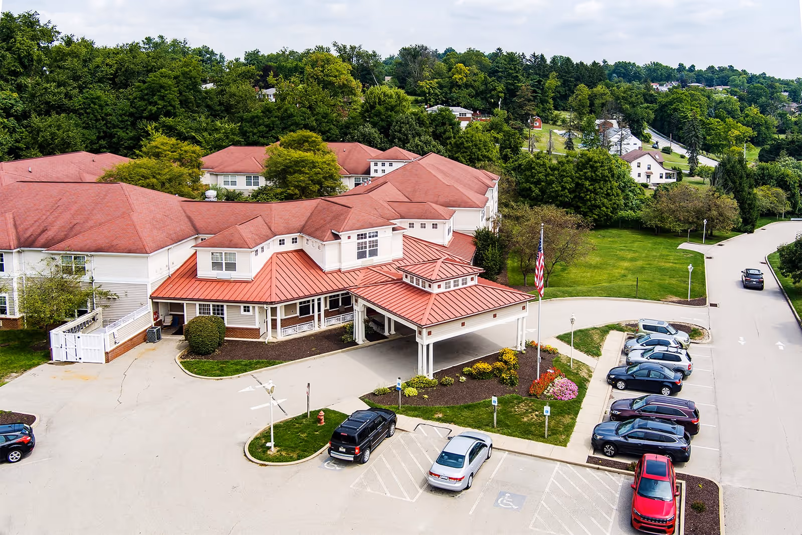 Aerial view of Hawthorne Woods by New Perspective senior living facility showing a large building with red roofs surrounded by greenery. There is a parking lot with several cars parked and a driveway leading to the entrance with a covered drop-off area. Trees and a grassy area are visible around the building.