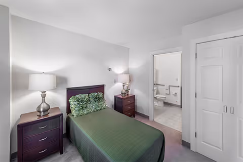 A small bedroom with a single bed covered in a green bedspread and two green pillows. There are two dark wood nightstands on either side of the bed, each with a lamp. The room has light-colored walls and carpeted floor. An open door reveals a bathroom with a toilet and grab bars.