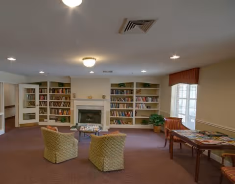 A cozy common room with two patterned armchairs facing a fireplace surrounded by built-in bookshelves filled with books. There is a table with chairs near a window with red valances, and a potted plant in the corner. The room has carpeted floors and ceiling lights.