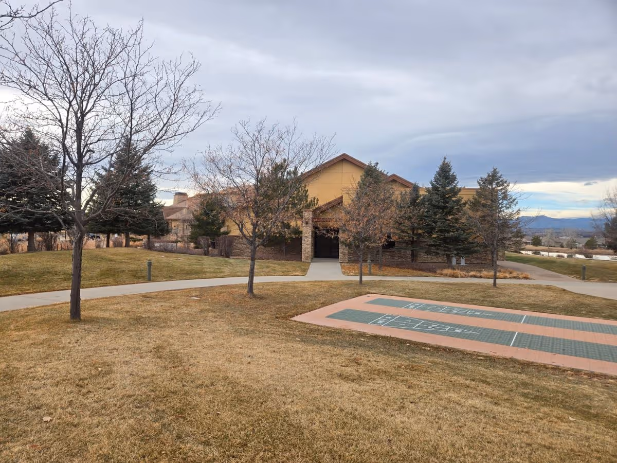 Front of a senior living facility building with a lawn, trees, walkways, and outdoor shuffleboard courts under a cloudy sky.