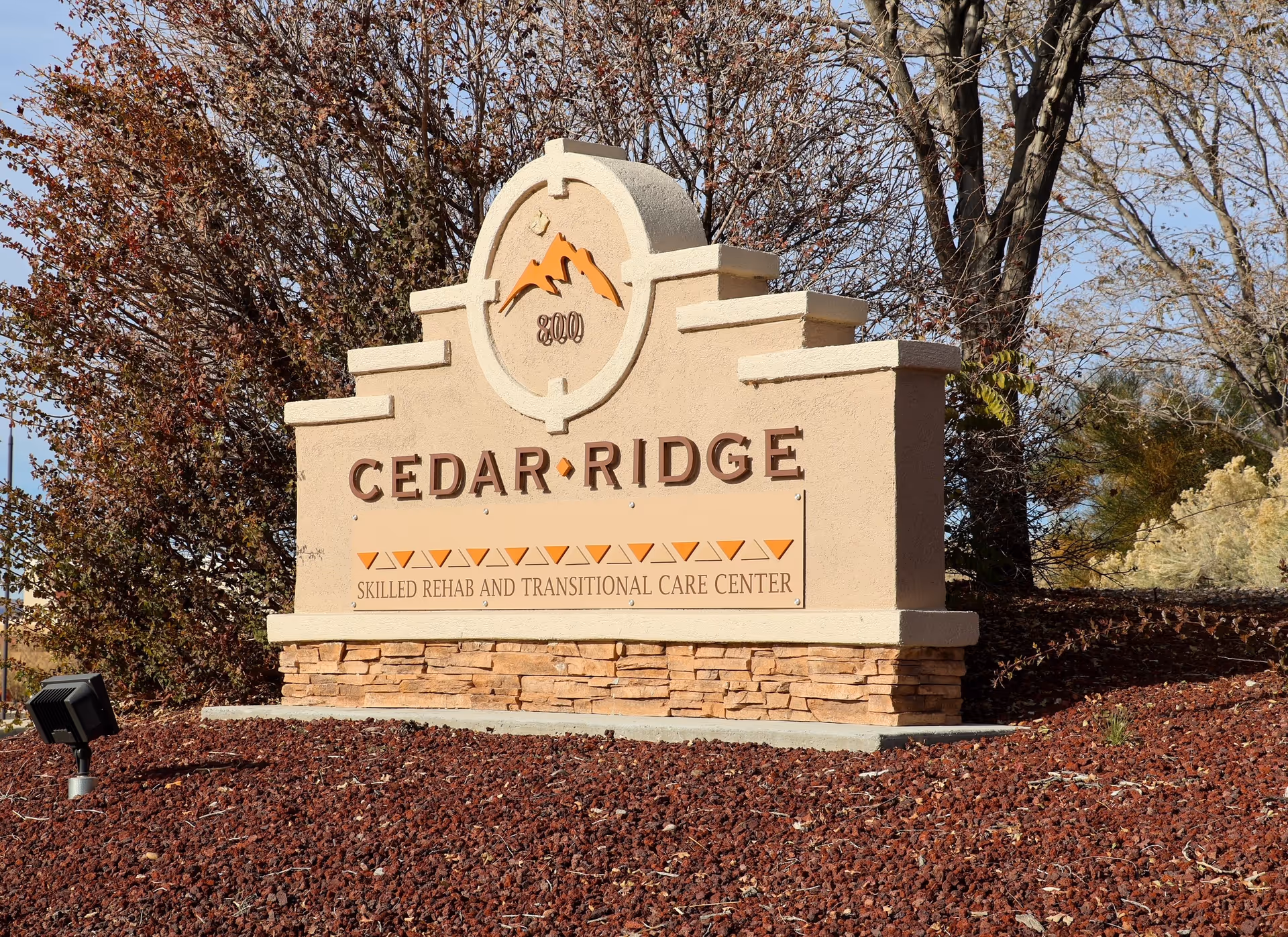 Outdoor stone sign for Cedar Ridge Skilled Rehab and Transitional Care Center surrounded by mulch and trees with autumn foliage.