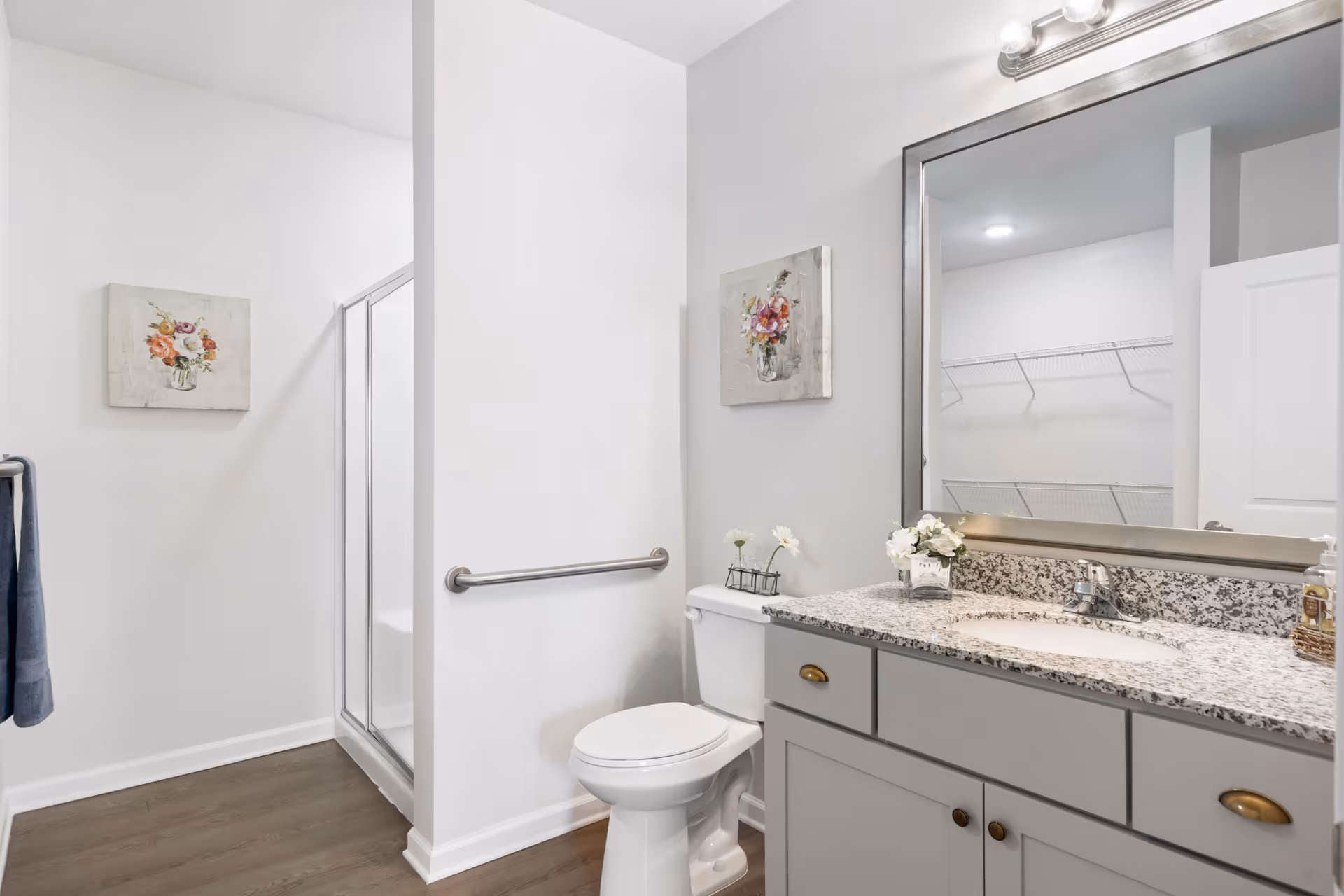 Bright and clean bathroom with a white toilet, granite countertop vanity with sink, large mirror, and a walk-in shower with glass door. The walls are white with a floral painting above the toilet and a towel hanging on a rack. The floor is wood-style.