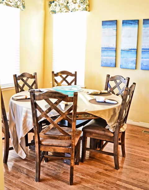 A dining room with a round wooden table covered by a beige tablecloth. Six wooden chairs with cushioned seats surround the table. The table is set with placemats and napkins. Two windows with floral valances allow natural light into the room. Three blue abstract paintings hang on the wall.