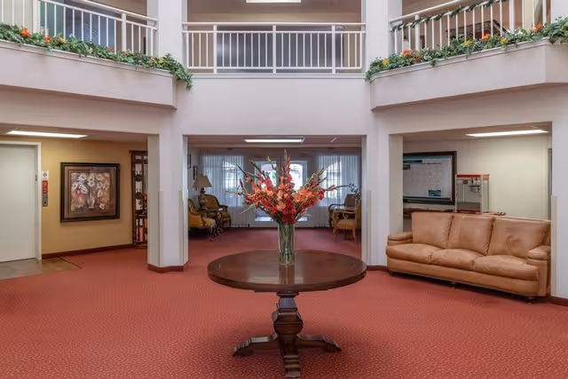 Interior view of a senior living facility lobby with a round wooden table in the center holding a vase of red flowers. The room has red carpet flooring, beige walls, and a beige couch on the right side. There are two seating areas with chairs and lamps in the background, and a second-floor balcony with white railings overlooking the lobby.
