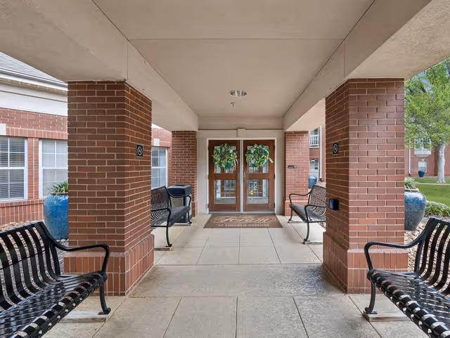 Covered entrance area with brick pillars and four black metal benches facing each other. Double glass doors decorated with green wreaths are at the end of the walkway. Large blue planters with greenery are placed near the pillars. The area is clean and well-maintained with a beige tiled floor.
