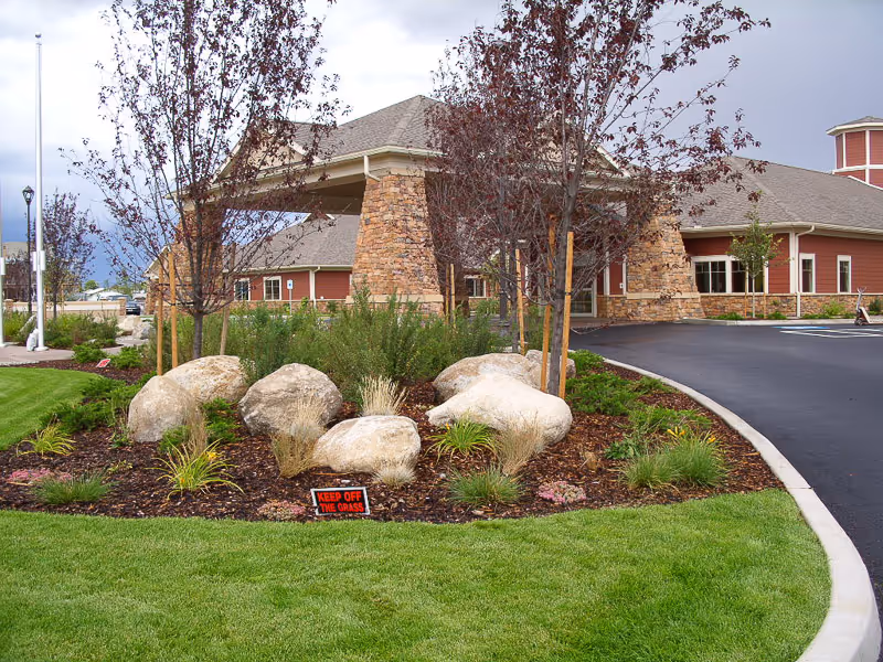 Entrance area of a senior living facility with a covered drop-off zone supported by stone pillars. The foreground features a landscaped garden with large rocks, small trees, and various plants, along with a 'KEEP OFF THE GRASS' sign. The building has red siding and a gray roof, and there is a paved driveway curving around the garden.