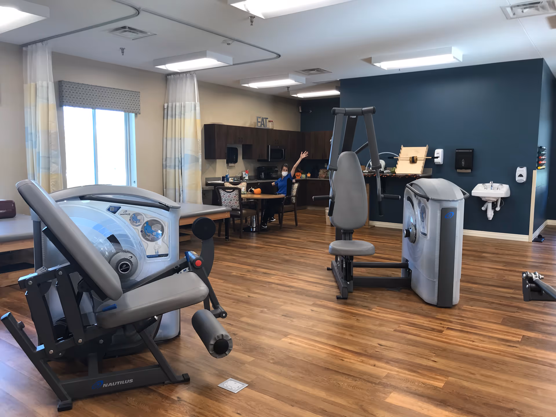 A spacious room with wooden flooring featuring exercise equipment including a leg press and a chest press machine. In the background, there is a kitchenette area with cabinets, a table, and chairs. A person wearing a mask is seated at the table waving. The room has large windows with curtains and a small wall-mounted sink.