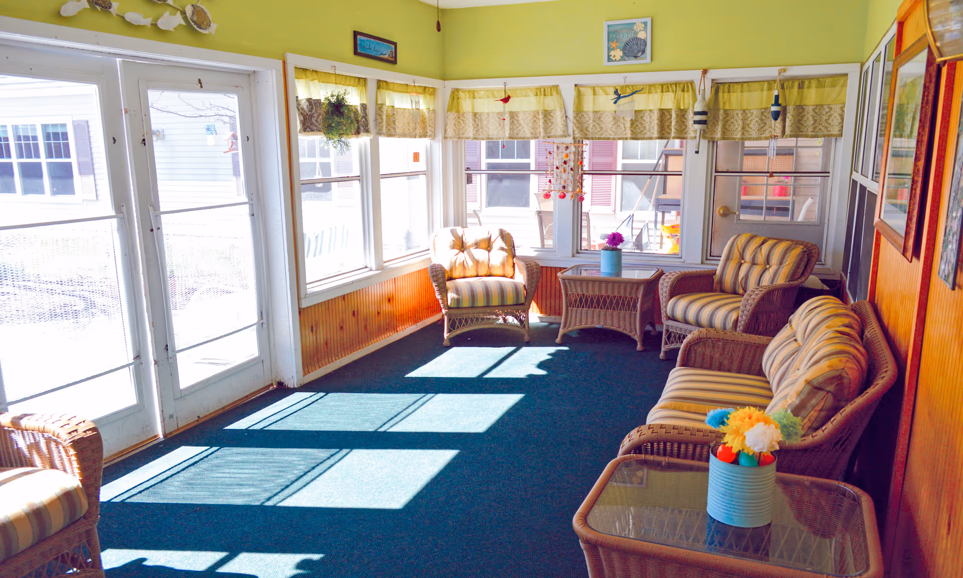 A sunlit sitting area with wicker furniture including a sofa, two armchairs, and two glass-top tables. The furniture has striped cushions. The room has large windows with yellow and beige valances, green walls, and a green carpet. There are decorative items hanging near the windows and small flower arrangements on the tables.