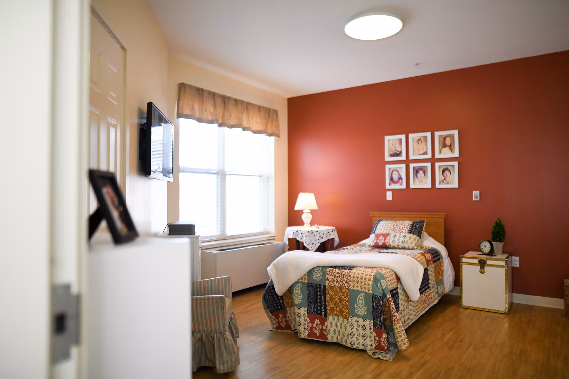 A cozy bedroom with a single bed covered in a colorful patchwork quilt. The bed is against a red accent wall decorated with six framed pictures. Next to the bed is a small table with a lamp and a white chest with a clock and a small plant on top. A window with blinds and a valance lets in natural light. A wall-mounted TV is visible on the left side of the room.