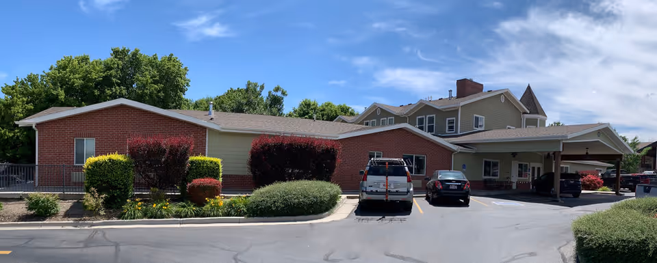 Exterior view of The Lodge at Jordan River, a single-story building with a brick and beige siding facade, surrounded by green shrubs and trees under a partly cloudy blue sky. Several cars are parked in front of the building near a covered entrance.