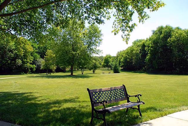 A peaceful outdoor scene at Geneva Lake Manor featuring a green grassy lawn with scattered trees and a black metal bench in the foreground. The sky is clear and blue, and the area is surrounded by dense green foliage.