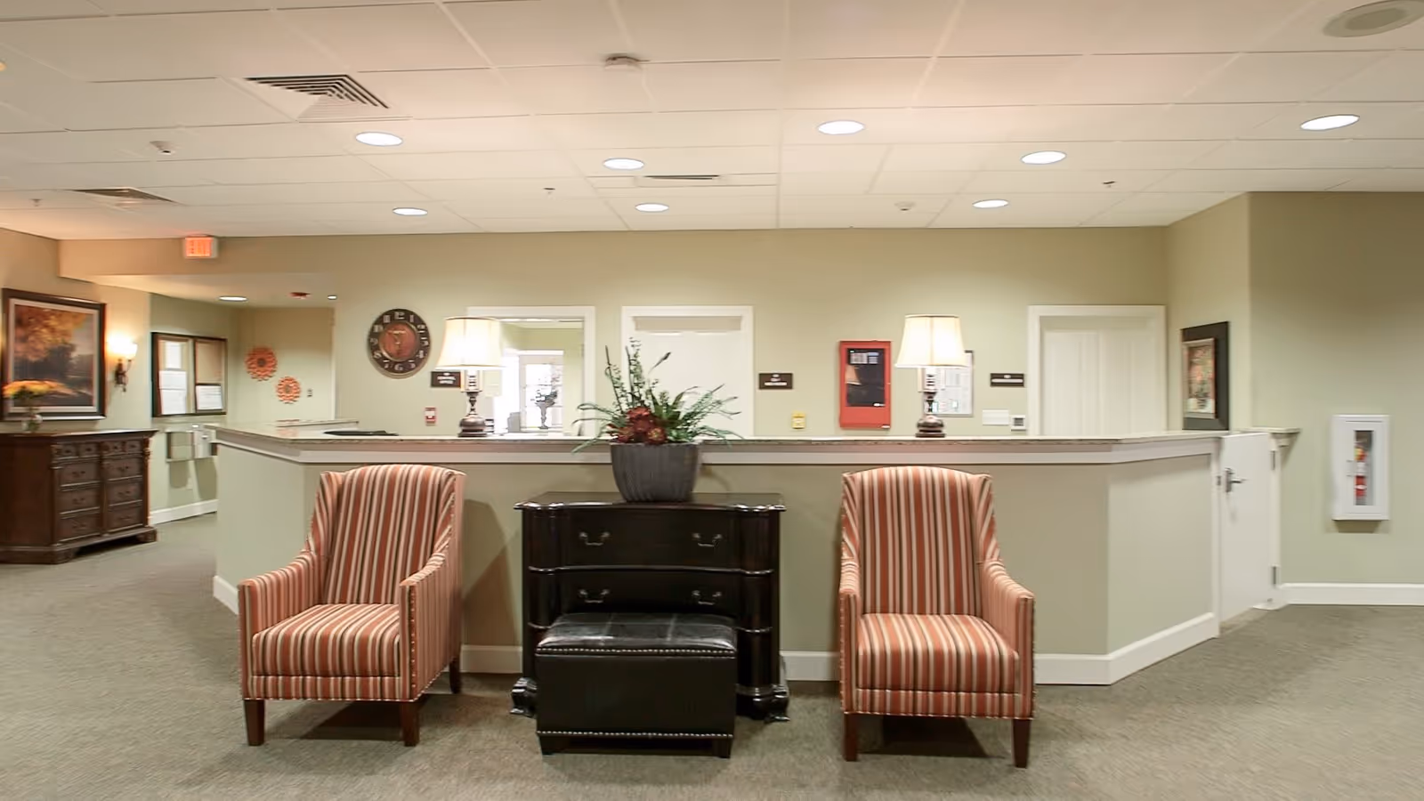 Interior view of a senior living facility reception area with two striped armchairs, a black chest of drawers with a plant on top, and two table lamps. The background shows a counter with three doorways, a clock, and a fire alarm panel on the wall.